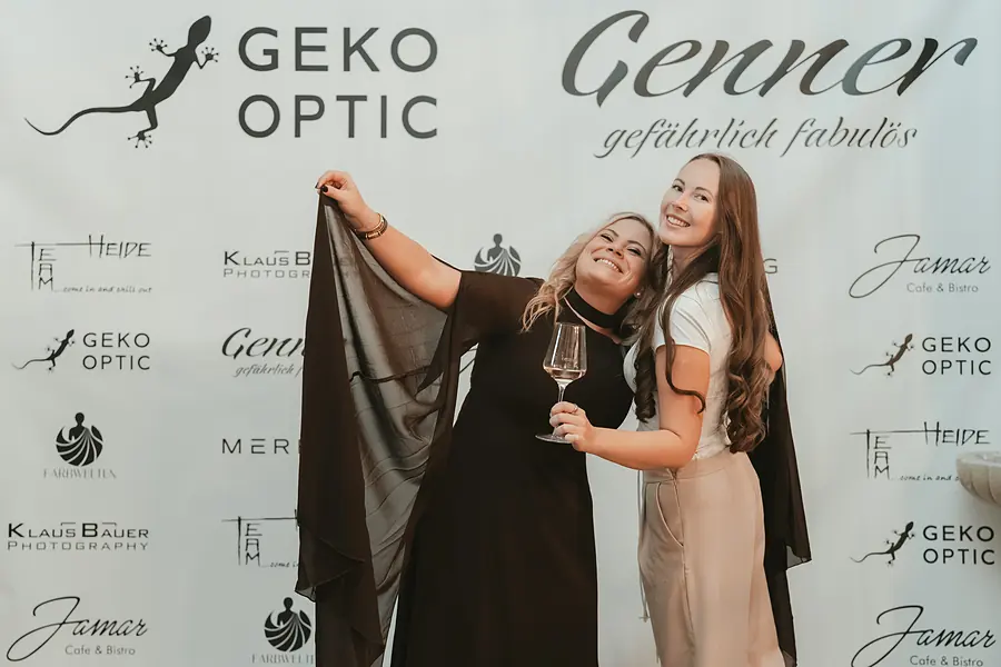 Zwei Gäste lachen vor der Fotowand, eine hält ein schwarzes Tuch hoch